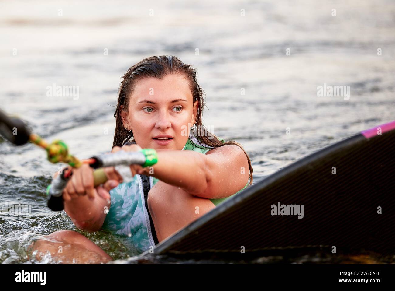 Giovane bella ragazza sportiva con un giubbotto di salvataggio verde seduta in acqua su un wakeboard con un cortile in mano. La sportiva si sta preparando per una stufa Foto Stock