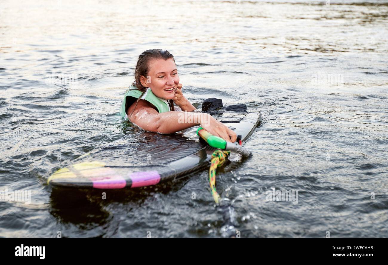 La giovane ragazza sportiva con un giubbotto di salvataggio verde nuota nell'acqua con il wakeboard e il cortile in mano. Felice sportiva che si prepara per una ripida Foto Stock