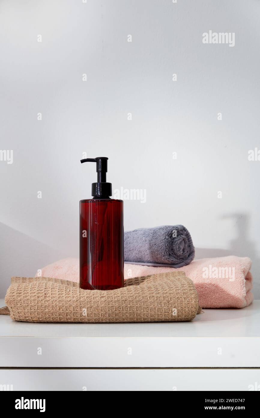 Immagine minima di un erogatore di bottiglie trasparente senza marchio posizionato su un asciugamano, decorato con altri asciugamani piegati. Concetto delicato per la cura della pelle Foto Stock