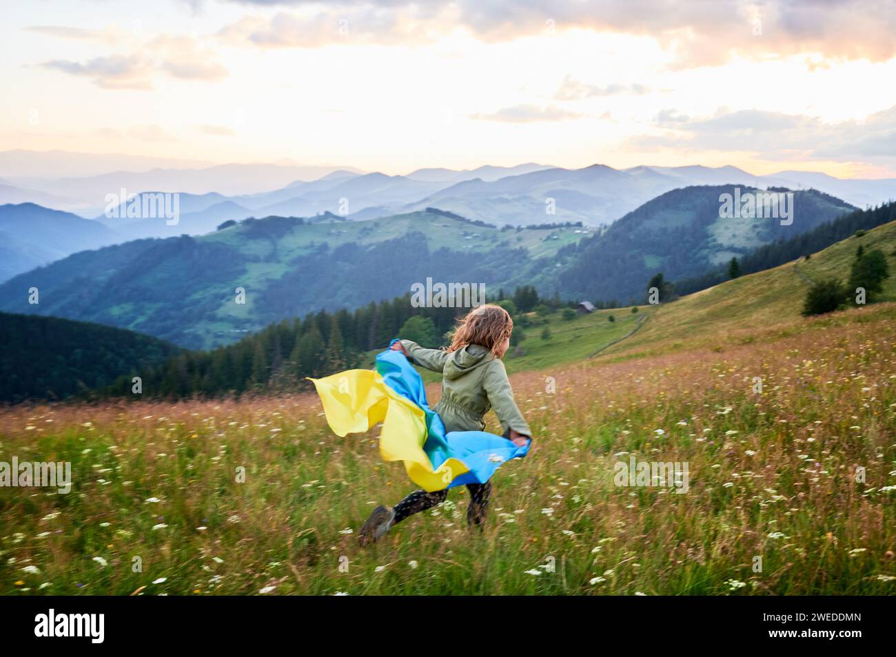 Ragazza felice che corre sulle colline con la bandiera Ucraina sulle spalle. Vista posteriore di bambina femmina flutting bandiera Ucraina. Piccolo bambino che corre sulle montagne dei Carpazi. Foto Stock
