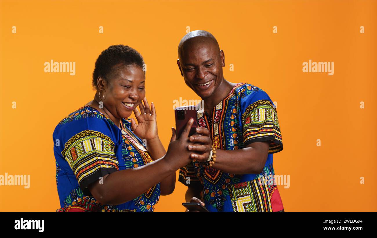 Coppia afroamericana tradizionale che naviga su siti Web da smartphone, controlla le foto sull'app dei social media e ridi di meme. Persone gioiose in abiti colorati che guardano le foto online. Foto Stock
