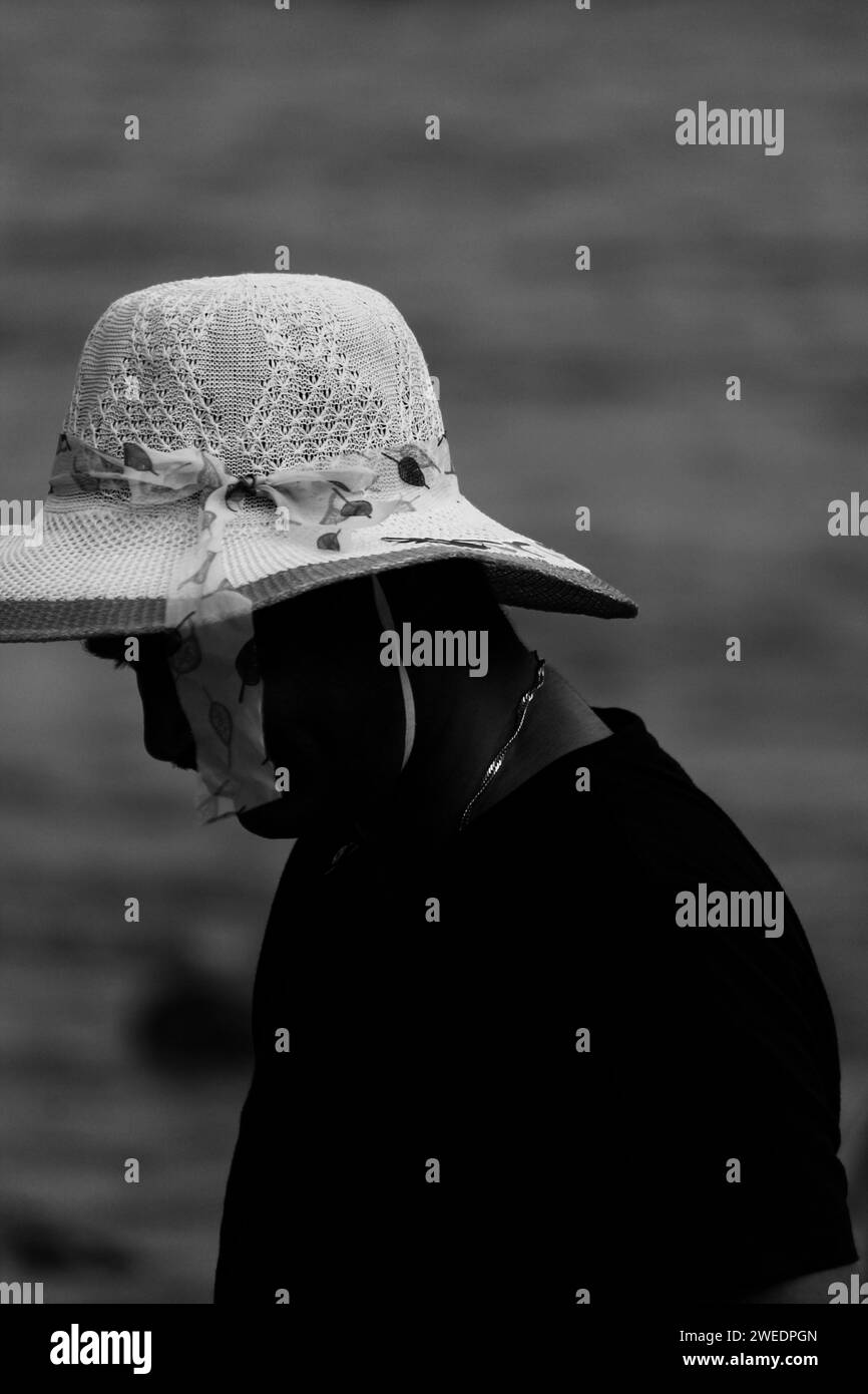 Una persona che guarda in basso nelle andamane con un cappello da donna. Bianco e nero. Spiaggia sullo sfondo Foto Stock