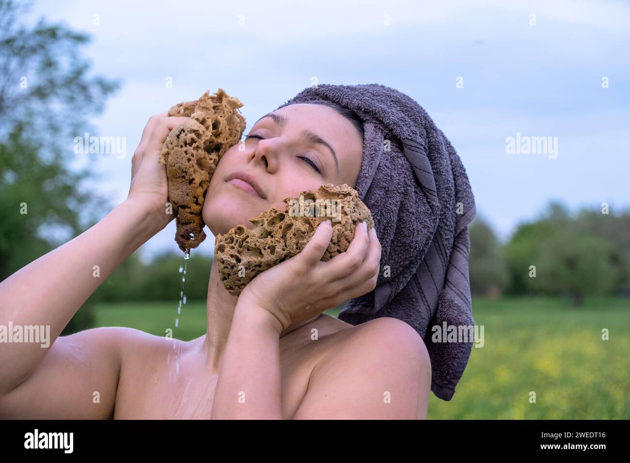 ritratto di una giovane donna nuda sexy e bella con la testa avvolta da un asciugamano, che fa un bagno nel verde prato della natura, lavandosi la testa con una spugna, copiando uno spazio Foto Stock