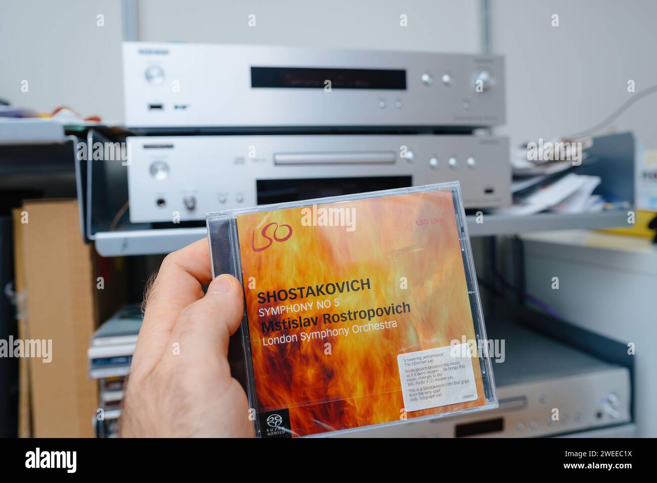 Parigi, Francia - 28 dicembre 2023: Male Hand Holding the SACD Super audio CD of Shostakovich's Symphony No. 5 diretto da Mstislav Rostropovich. Sullo sfondo, un impianto hi-fi e un deck in argento SACD completano la configurazione per audiofili Foto Stock