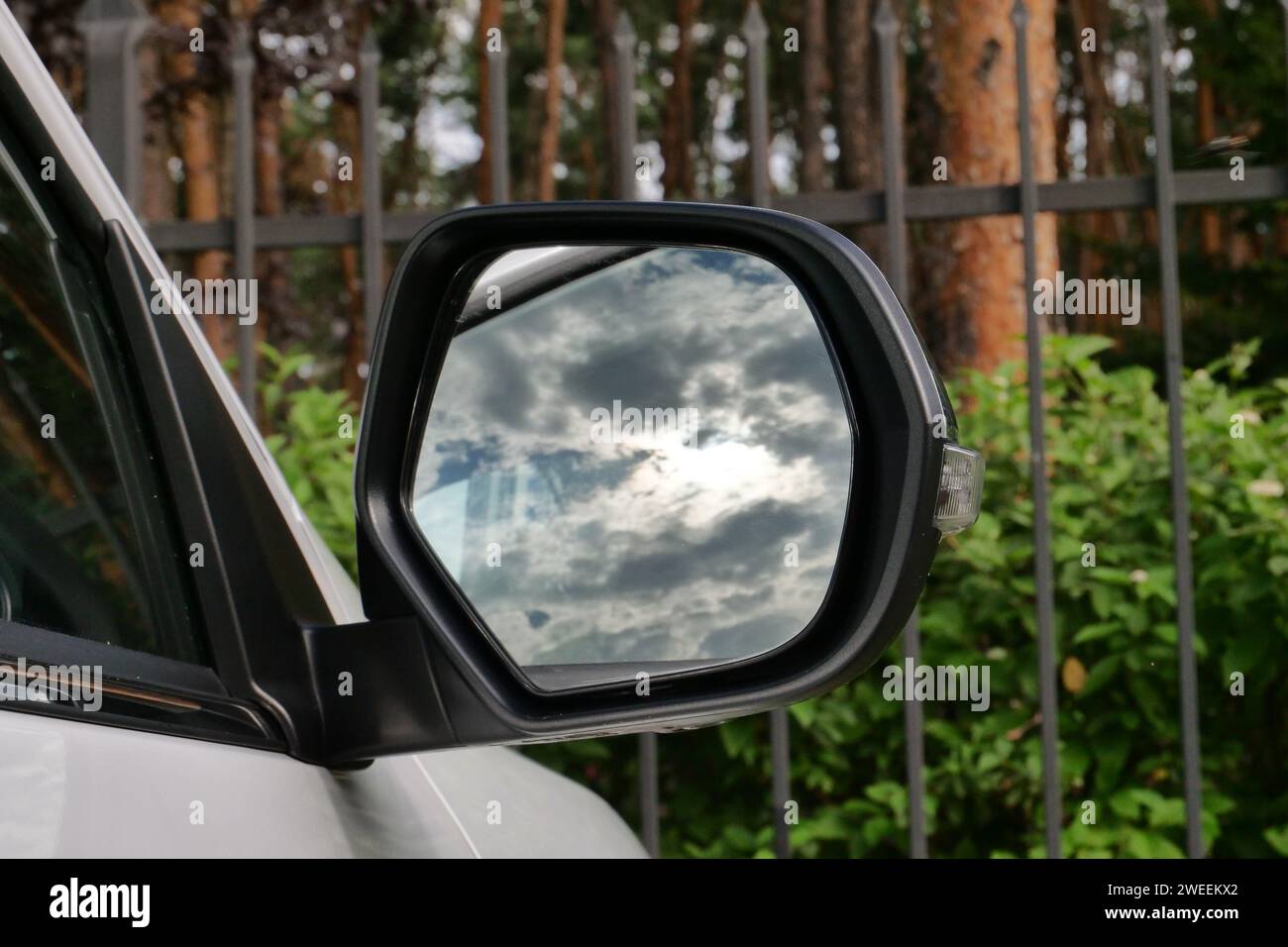 Il cielo nuvoloso si riflette nello specchio di un'auto parcheggiata vicino alla recinzione di ferro Foto Stock