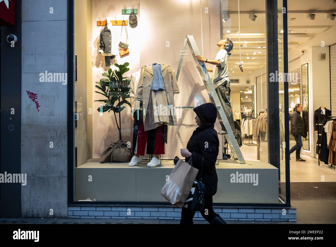 Una donna si trova su una scala a pioli all'interno di un negozio di abbigliamento mentre riorganizza la vetrina in Oxford Street, Londra, Inghilterra, Regno Unito Foto Stock