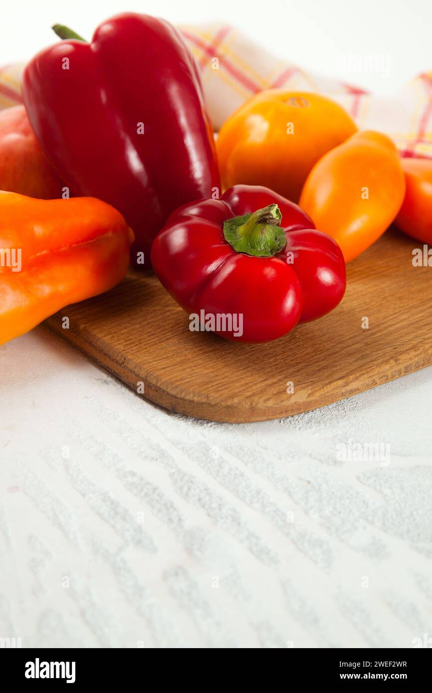 Immagine di sfondo vegetale. Tagliere con assort di diversi pomodori succosi interi e peperoni maturi su fondo di legno bianco con kitch rosso Foto Stock
