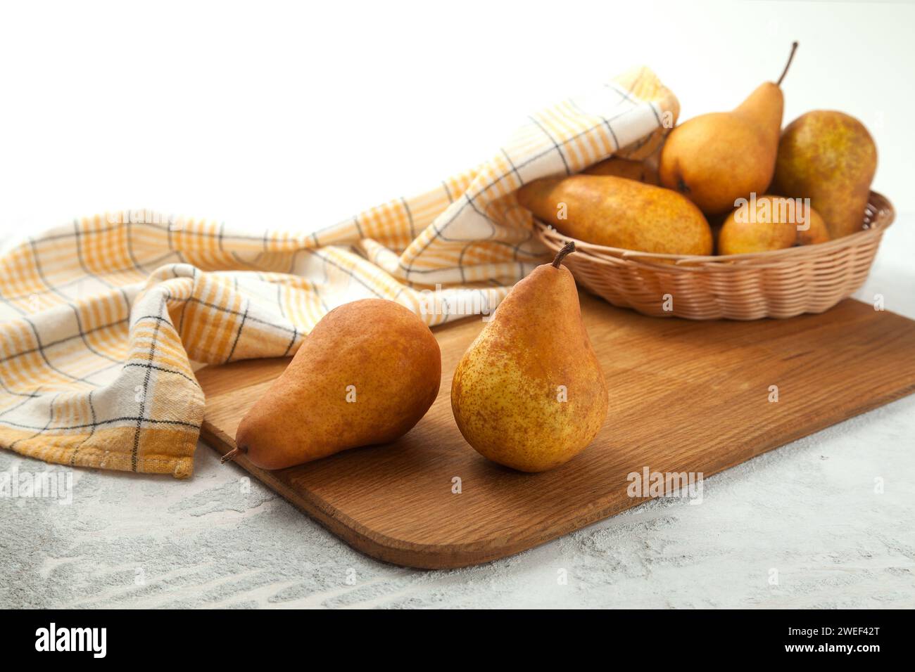 Tagliere in legno con frutta di pera matura e succosa, cesto di vimini con pere e asciugamano da cucina giallo su fondo di legno bianco. Foto Stock