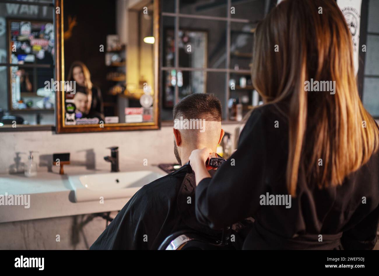 Giovane barbiera che crea un taglio di capelli tagliati da uomo utilizzando il tagliacconciatura di fronte allo specchio barbiere in stile moderno. Servizio di cura dei capelli sma locale Foto Stock