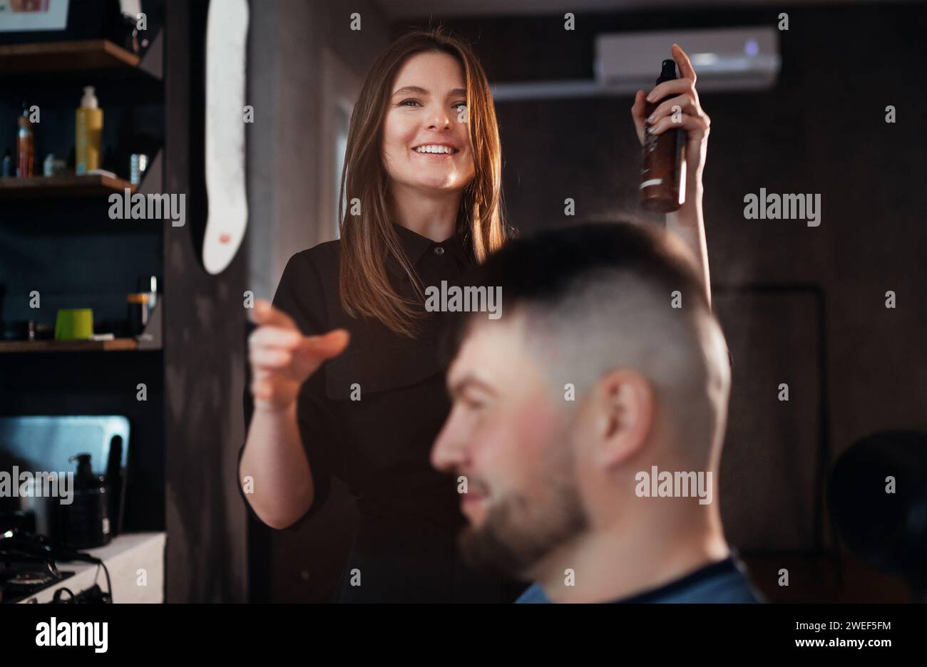 Sorridente giovane parrucchiere che spruzza profumo eau de toilette dopo l'acconciatura di un uomo barbuto. Interni moderni in stile barbiere nero chiaro. Ha Foto Stock