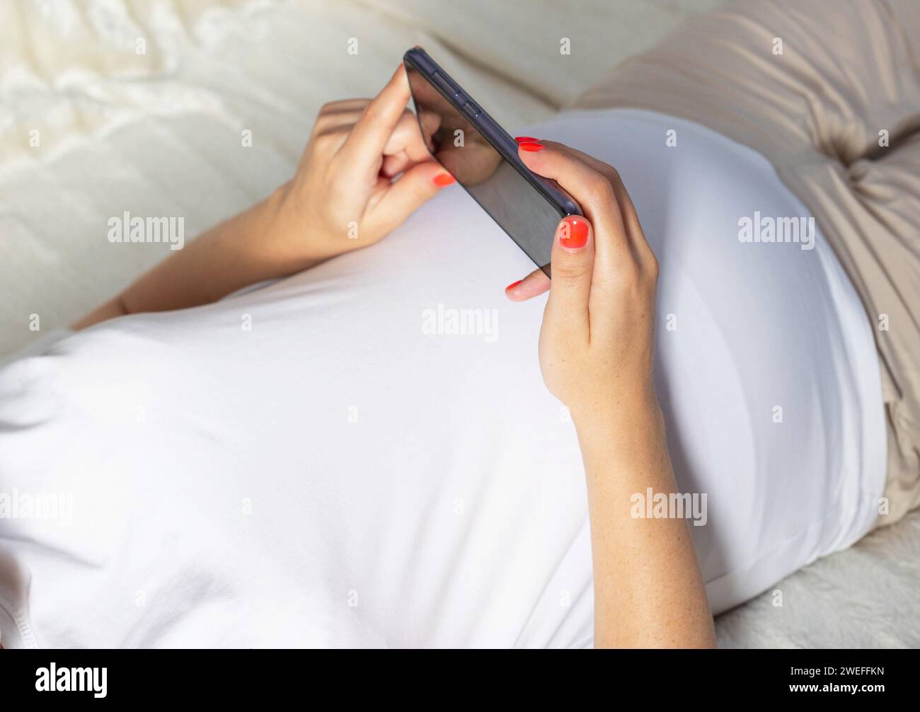 Una ragazza incinta è sdraiata sul letto e tiene uno smartphone tra le mani. Concetto di applicazioni negli smartphone per donne in gravidanza, numero di contrazioni Foto Stock