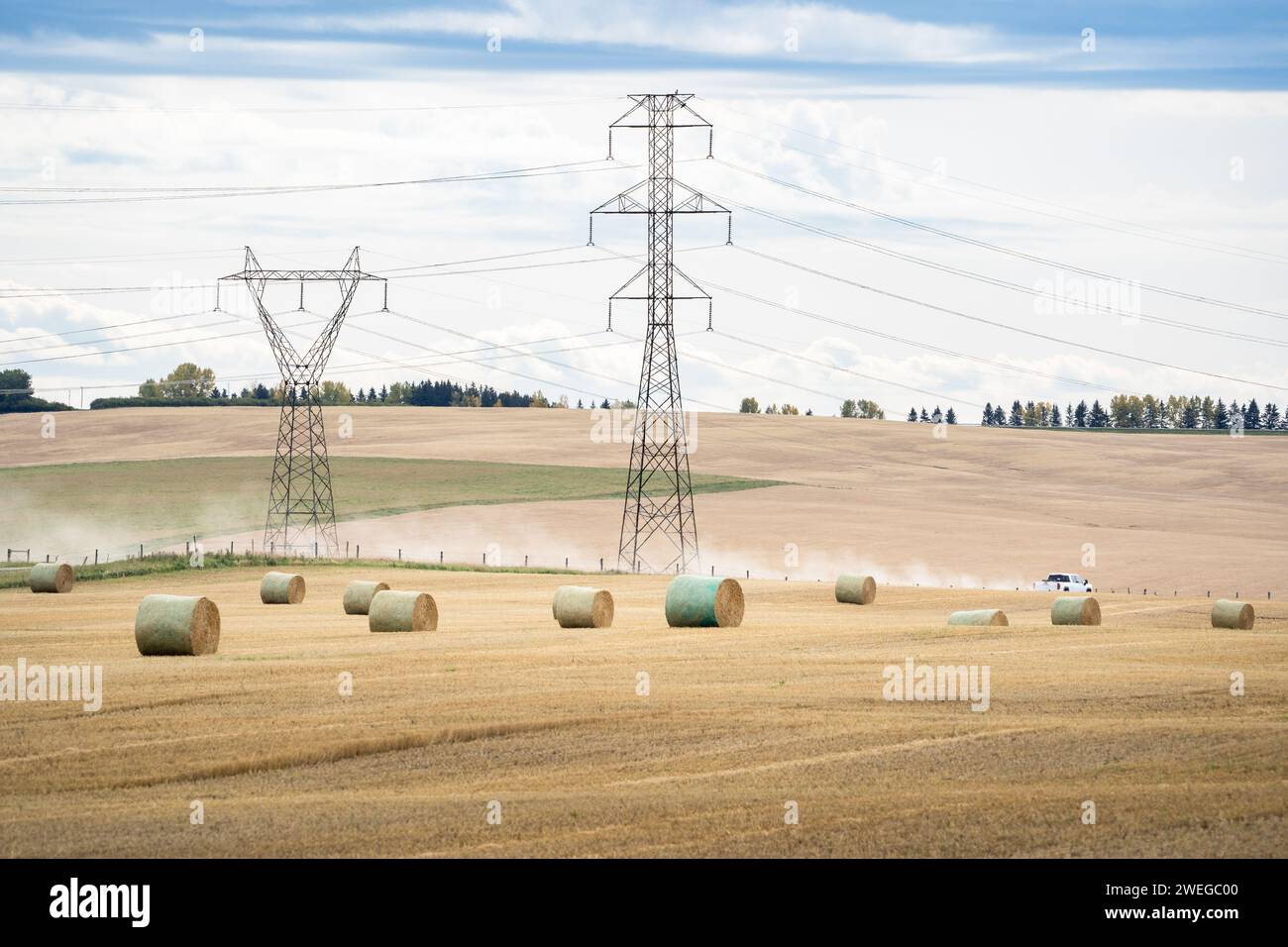 Rotoli rotondi su un campo di raccolta che si affaccia su piloni elettrici distanti delle aziende di servizi pubblici nelle praterie dell'Alberta con un camion e una pista di polveri su una tomba Foto Stock