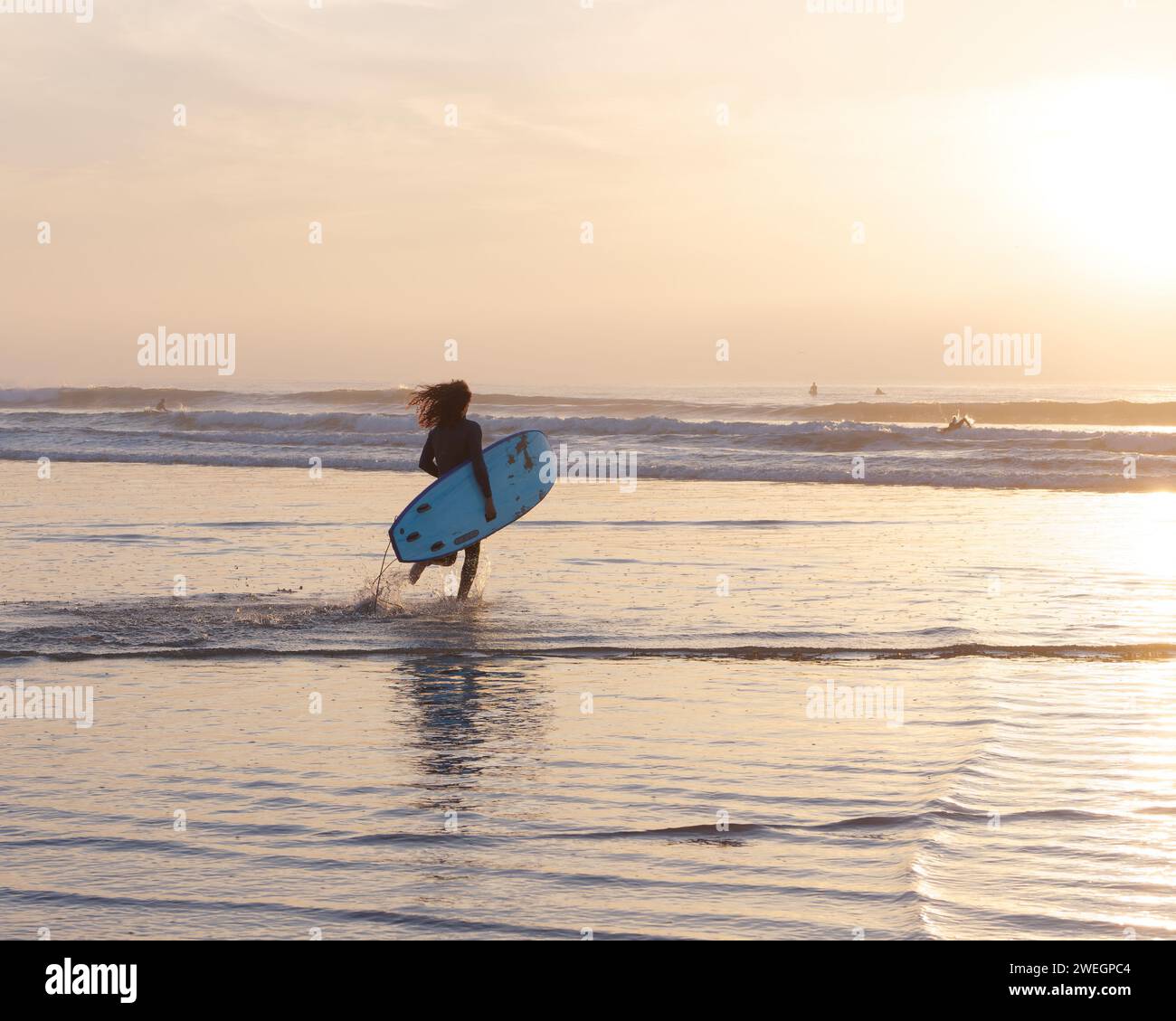Surfista maschile in muta con lunghi capelli corre verso l'acqua mentre sta per fare surf al tramonto a Essaouira, Marocco, 25 gennaio 2024 Foto Stock