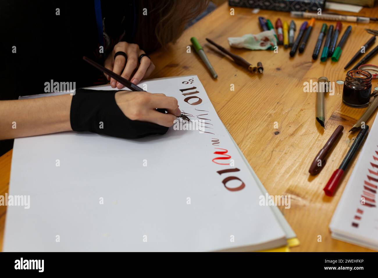 Una calligrafa esperta pratica a scrivere e disegnare caratteri in stile rinascimentale in un quaderno di schizzi. Sul tavolo di legno sono presenti marcatori e colori Foto Stock