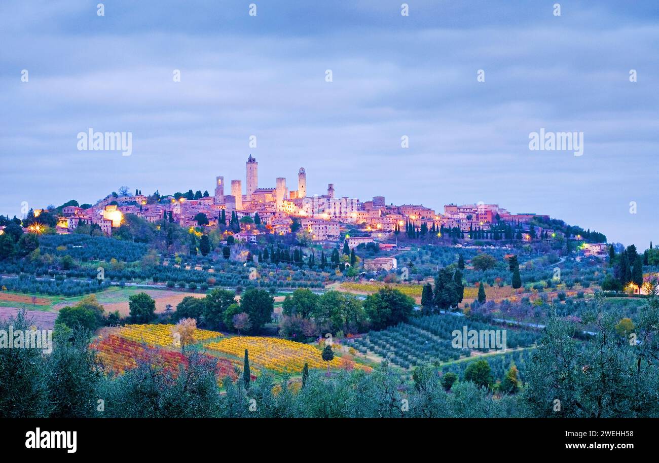Vista sulla città di San Gimignano su una collina la mattina presto all'ora blu con cielo nuvoloso in autunno, cipressi, oliveti e vigneti, Foto Stock