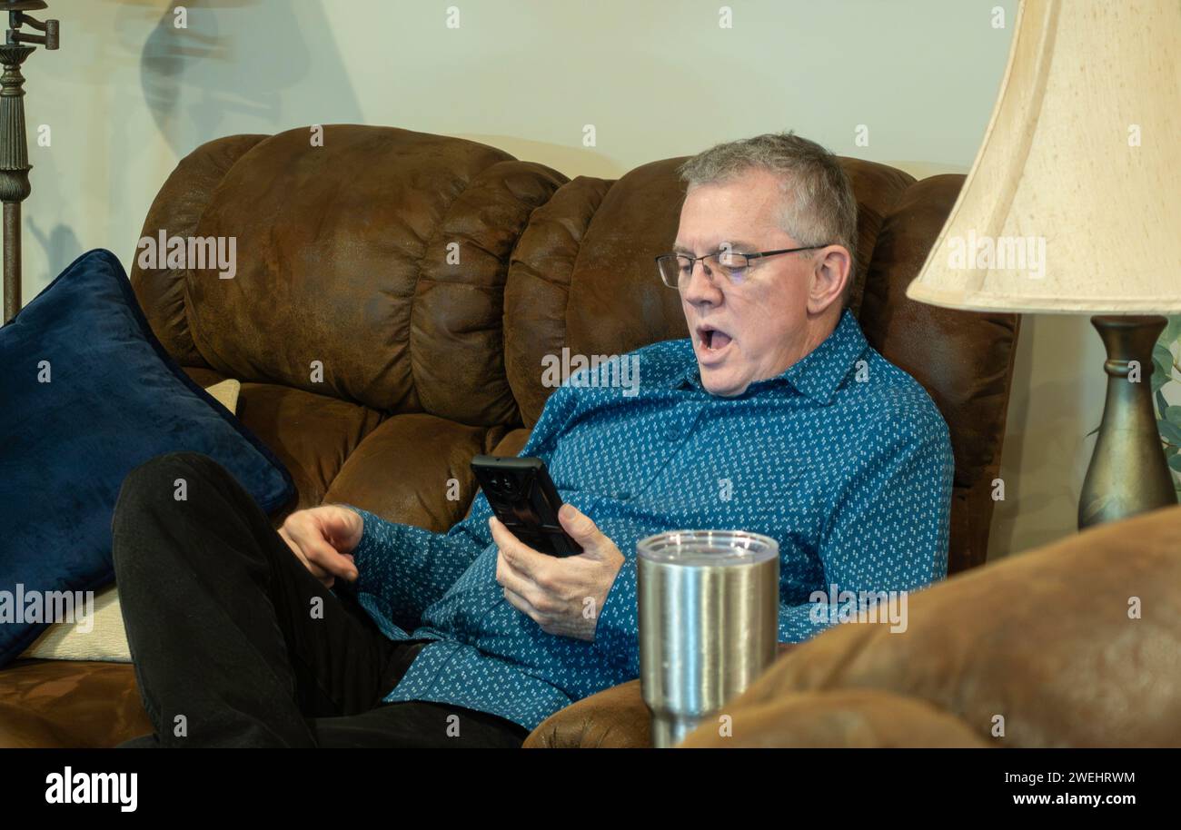 Uomo caucasico adulto di 60 anni che si rilassa sul divano e fissa qualcosa sul telefono con shock o sorpresa. Foto Stock