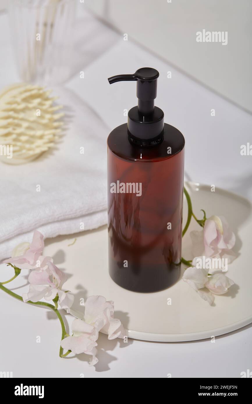 Un biberon ambrato senza etichetta con bocchetta, mockup per prodotti per la cura dei capelli su vassoio rotondo, fiori, spazzola massaggiante per cuoio capelluto e asciugamano di cotone decorato su di esso Foto Stock