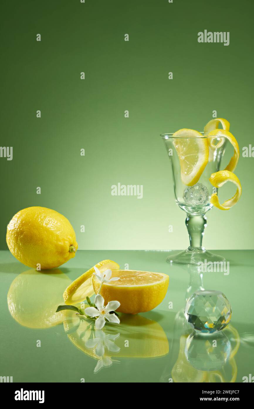Limone fresco, fette e buccia di limone decorata con tazza di vetro e palla di cristallo su sfondo verde retroilluminato. Concetto minimale, spazio per prodotti cosmetici Foto Stock