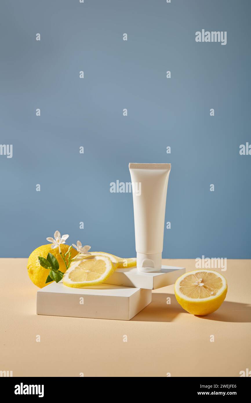 Scenografia minima per la pubblicità: Tubo di plastica bianco su podi, fette e fiori di limone fresco su sfondo blu. Mockup per prodotti cosmetici di lem Foto Stock