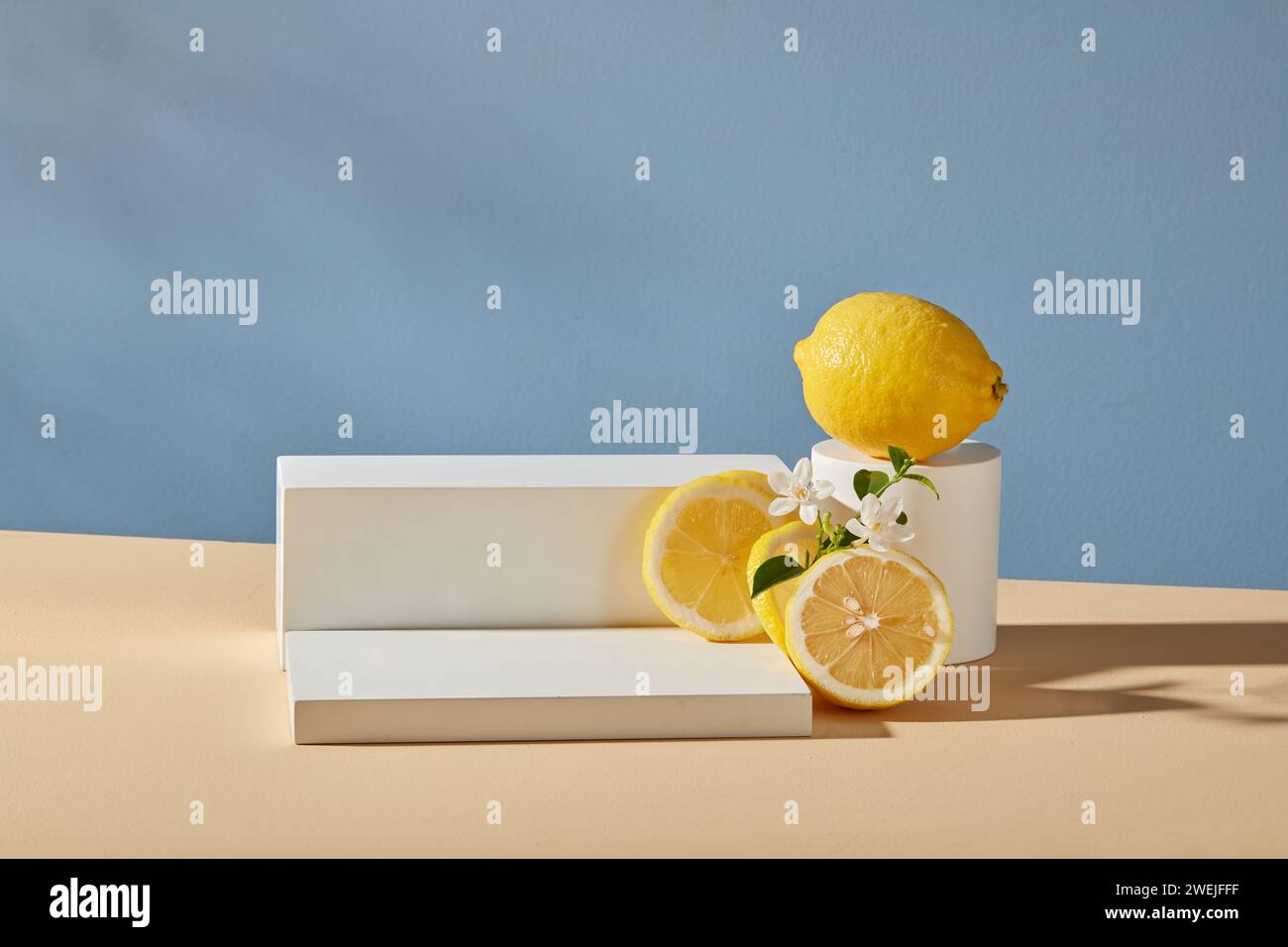 Scena minimalista per l'esposizione di cosmetici: Podi bianchi su sfondo blu con limone fresco, fette e fiori. Il limone contiene molta vitamina C che funziona Foto Stock