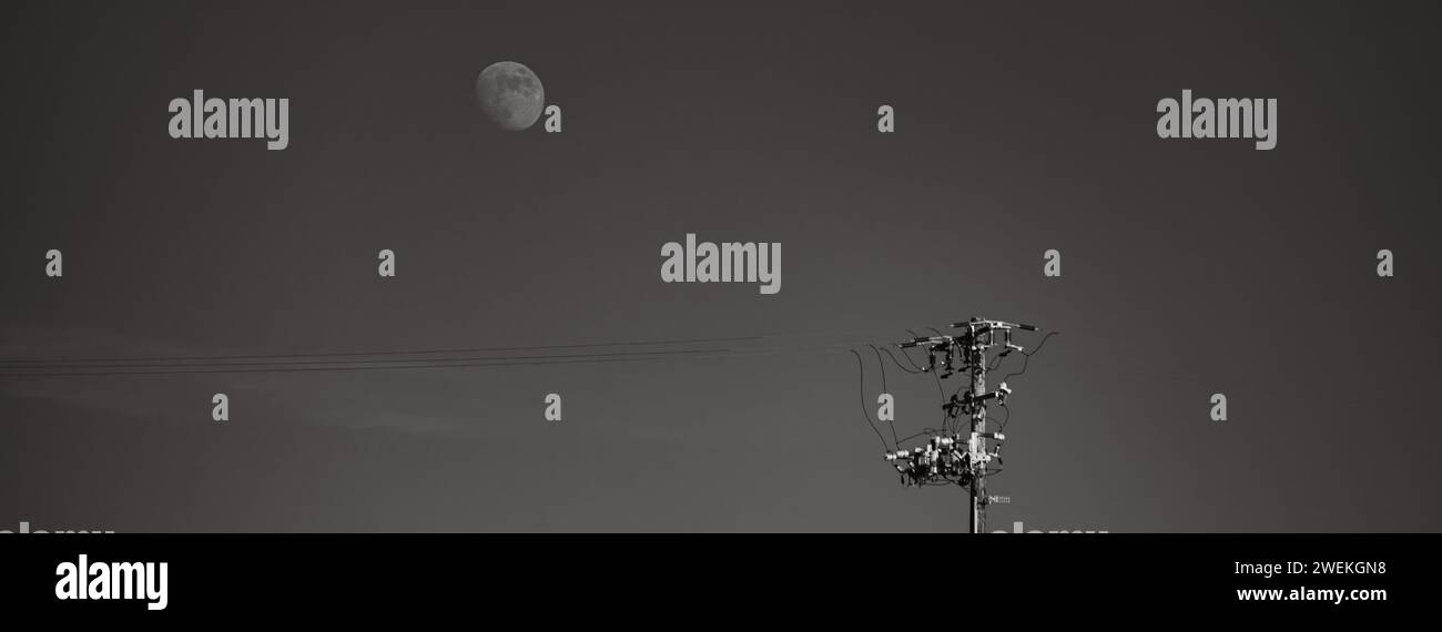 Uno scatto in bianco e nero di un palo elettrico contro un cielo notturno con una luna prominente. Foto Stock