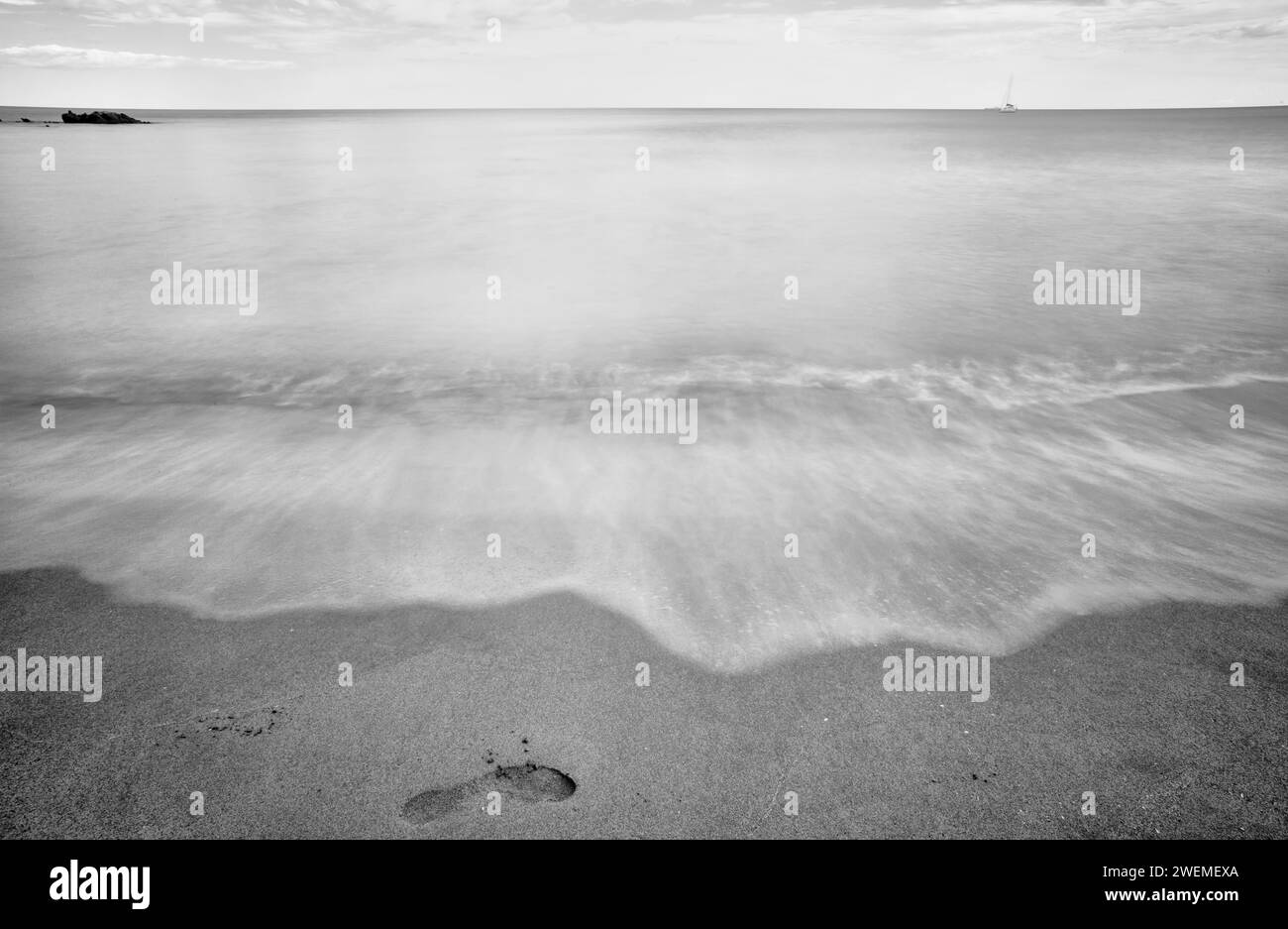 Serena esposizione bianca e nera di una tranquilla spiaggia sarda con una barca a vela all'orizzonte Foto Stock