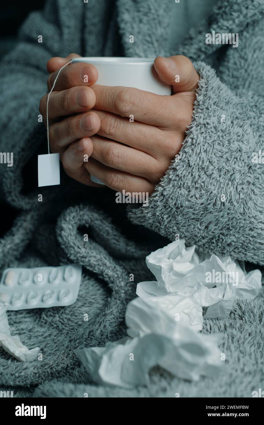 primo piano di un uomo malato a casa, in un abito da casa grigio, circondato da tessuti usati, che si riscalda con una tazza di tè caldo alle erbe Foto Stock