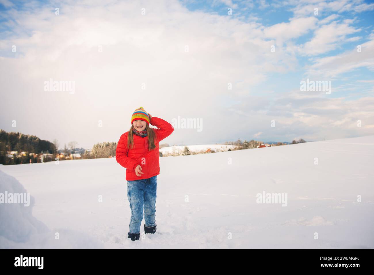 Bambina che indossa una giacca rossa e un cappello colorato, che gioca con la neve in inverno Foto Stock