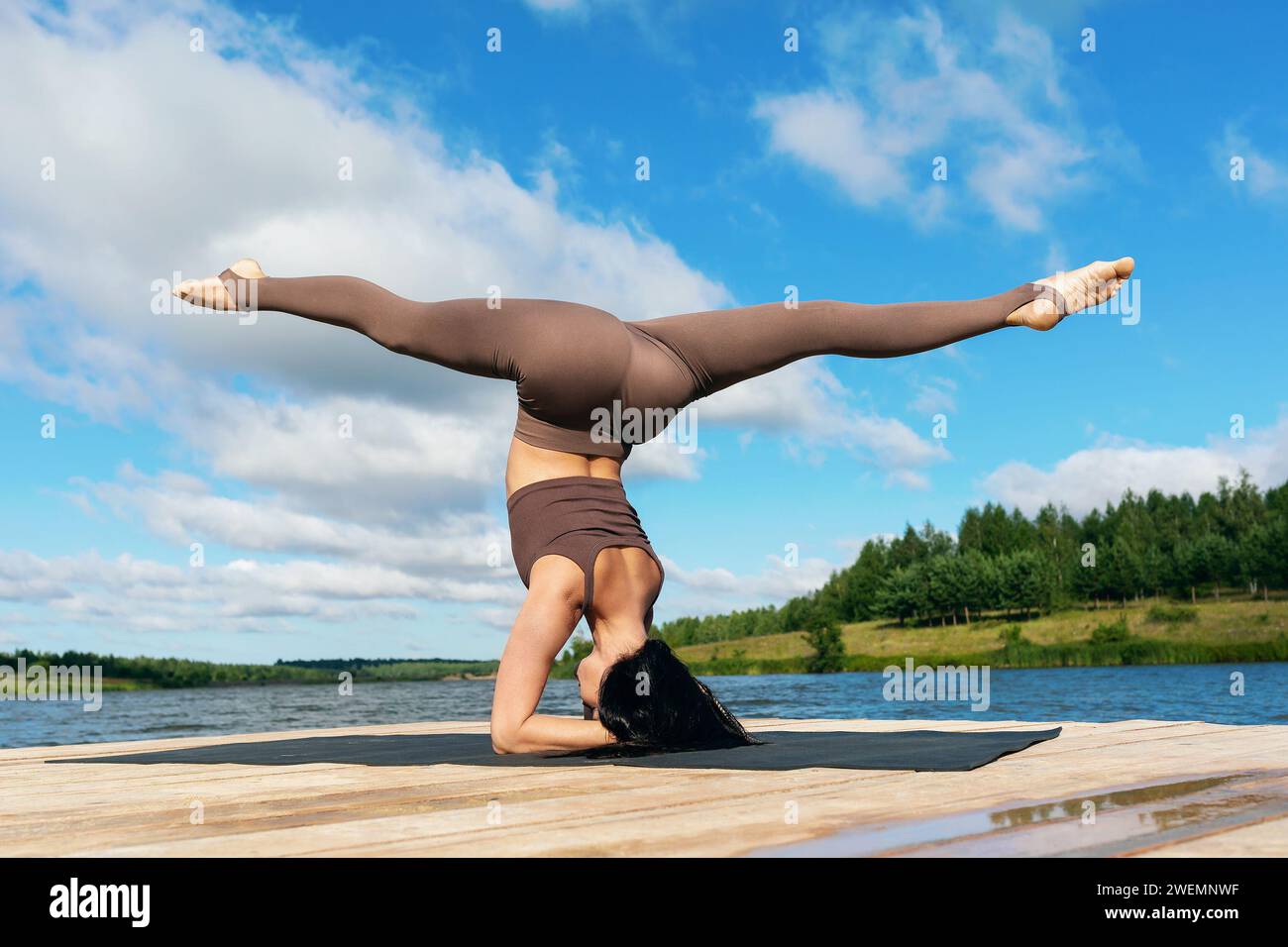 Donna attraente che esegue l'esercizio Shirsasana con Hanumanasana, asana invertita, poggiatesta con spaccature longitudinali, allenamento sulla riva di un lago Foto Stock