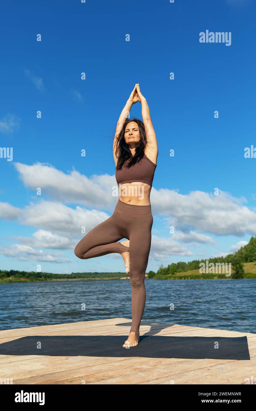 Donna attraente che esegue l'esercizio Vrikshasana, posa degli alberi, in piedi con le braccia sollevate e unite insieme, che si allenano con leggings e una maglia corta Foto Stock