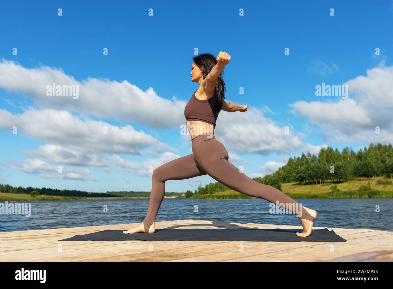 Donna attraente che esegue una variante di esercizio Virabhadrasana, posa guerriera, allenamento con leggings e una maglia corta mentre si trova sulla riva di Foto Stock