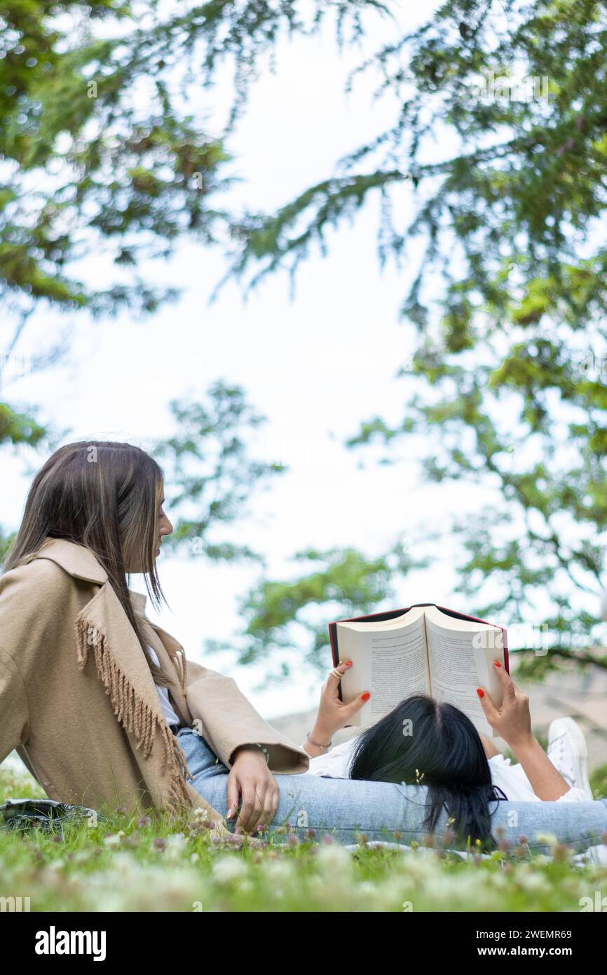 Vista laterale di due amiche rilassate che si godono una giornata insieme nel parco mentre prendono un po' d'aria fresca e leggono un libro Foto Stock