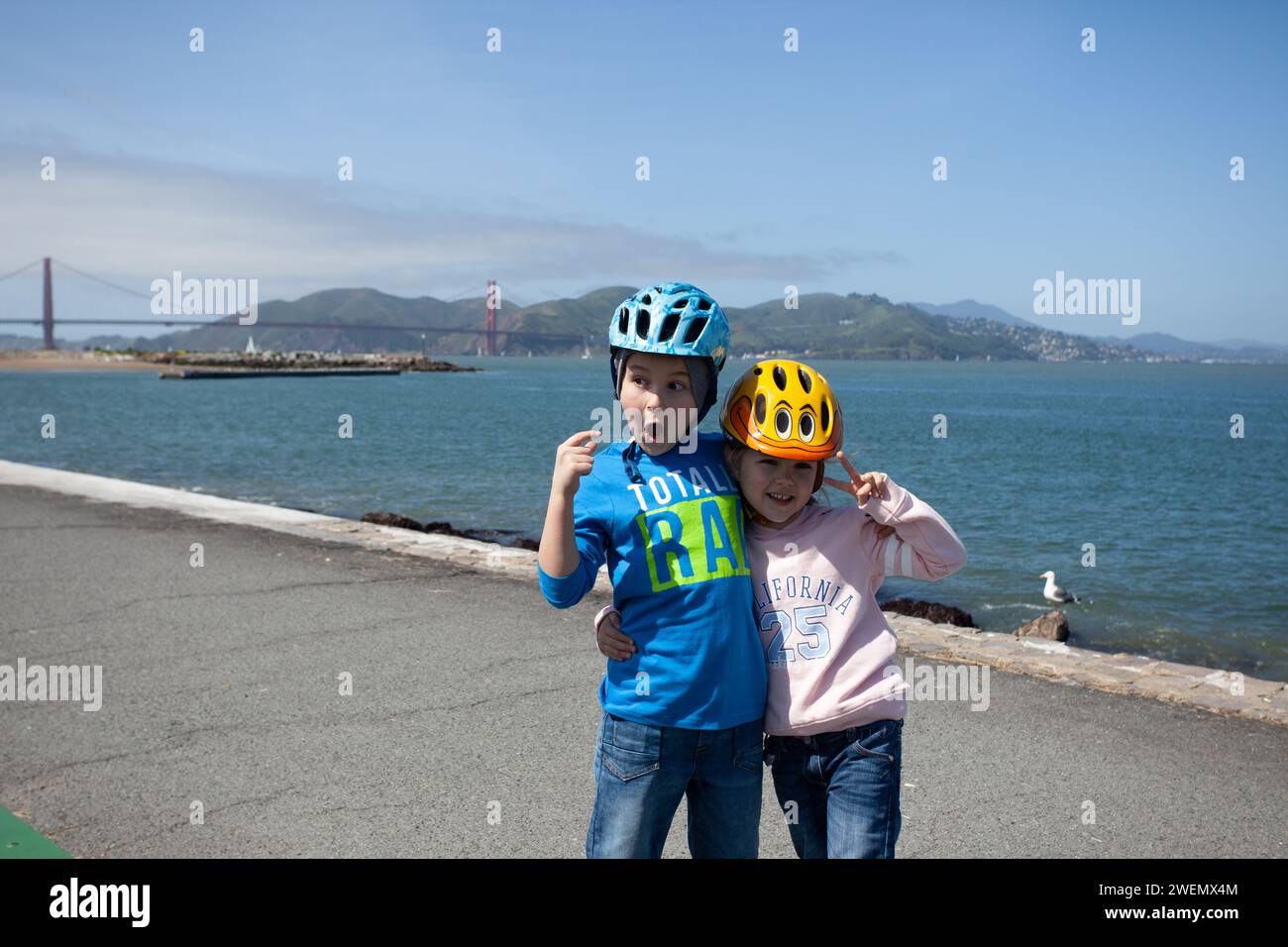 Un giovane ragazzo e una giovane ragazza sorpresi dal Golden Gate Bridge di San Francisco, che indossa jeans e caschi da bicicletta con uno sguardo sorpreso sul viso Foto Stock