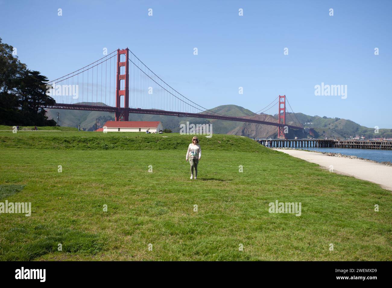 Il Golden Gate Bridge è un ponte sospeso che attraversa il Golden Gate, con una giovane donna che cammina sotto di esso sull'erba con il ponte in lontananza. Foto Stock