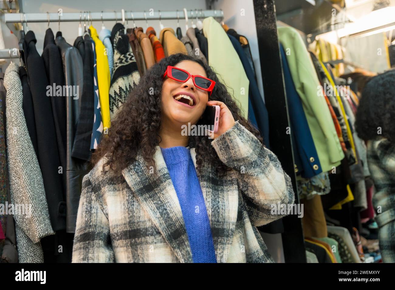 Giovane donna elegante e divertente che parla con il cellulare all'interno di un negozio pieno di vestiti in vendita Foto Stock