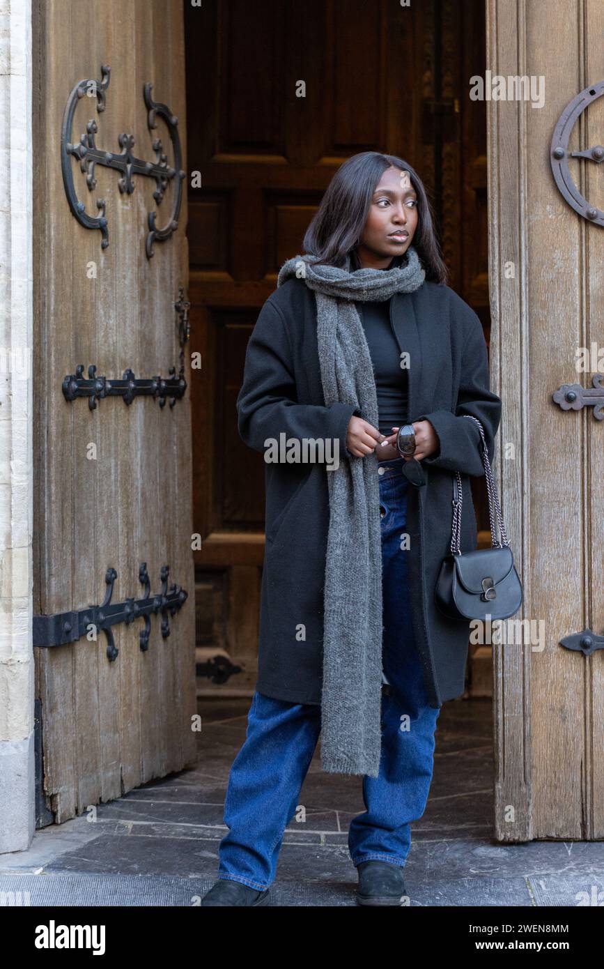Catturata all'ingresso di una cattedrale gotica, una giovane donna si trova in contemplazione, il suo sguardo è diretto verso il mondo esterno. Il significato storico della località è palpabile, con le elaborate cerniere delle porte e le lavorazioni in pietra che raccontano secoli di storia. Il suo abbigliamento, un mix di modernità chic e calore funzionale, suggerisce una giornata trascorsa alla ricerca della scoperta tra i sussurri del passato. Bellezza contemplativa all'ingresso della cattedrale. Foto di alta qualità Foto Stock