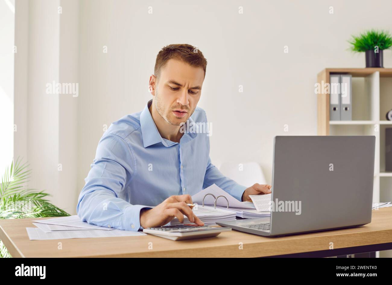 Commercialista di sesso maschile che lavora in ufficio, fa documenti cartacei e utilizza un computer portatile con calcolatrice Foto Stock