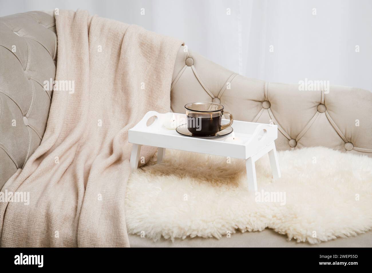 Vassoio per la colazione in legno bianco con le gambe sul divano di casa con una tazza di caffè caldo e candele in fiamme. Allestimento Hygge con coperta a quadri di lana morbida. Foto Stock