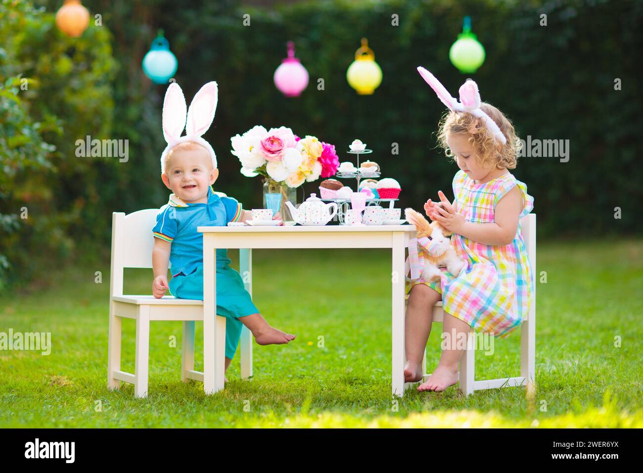 Bambini con la caccia all'uovo di Pasqua. Bambino e ragazza che indossa orecchie da coniglio che gioca a un tè, mangiando uova di cioccolato e torta nel soleggiato giorno di primavera. Foto Stock