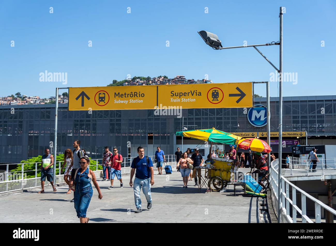 Pedoni che lasciano la stazione ferroviaria di Maracana, passando accanto a un cartello con le informazioni sulla stazione, sopra la passerella sotto il cielo azzurro della mattina d'estate. Foto Stock