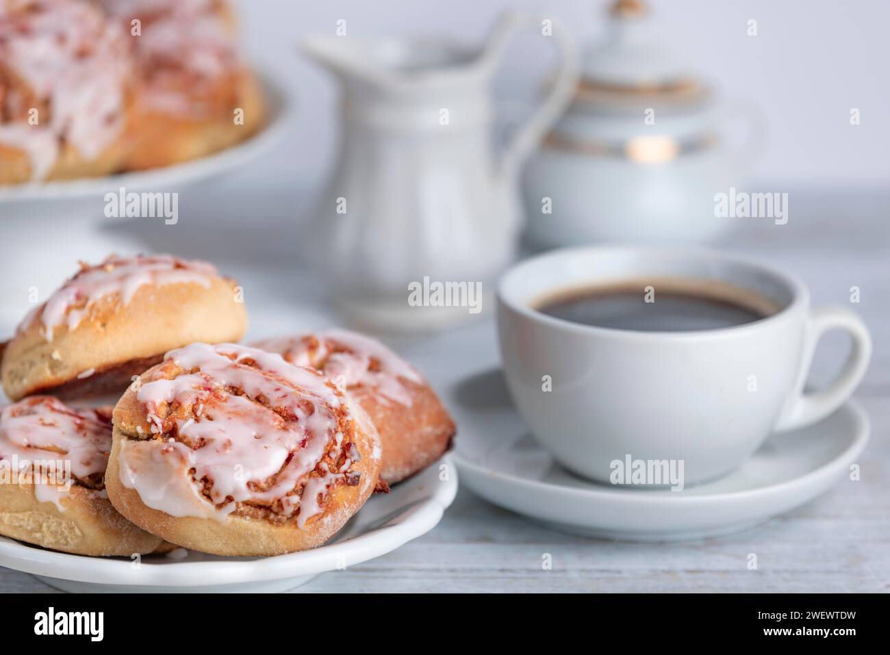 Pausa caffè con panini alla cannella accanto a una tazza di caffè, brocca per la panna e ciotola per lo zucchero sullo sfondo Foto Stock