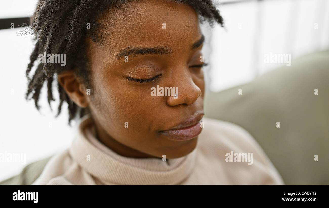 Giovane donna afroamericana con i dreadlock in una stanza dai toni neutri, occhi chiusi, che riflette la tranquillità. Foto Stock