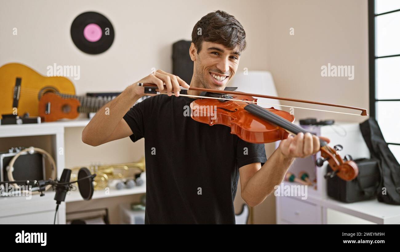 Giovane artista ispanico sicuro e sorridente che suona il violino felicemente in uno studio di musica al coperto, una sinfonia di intrattenimento classico Foto Stock