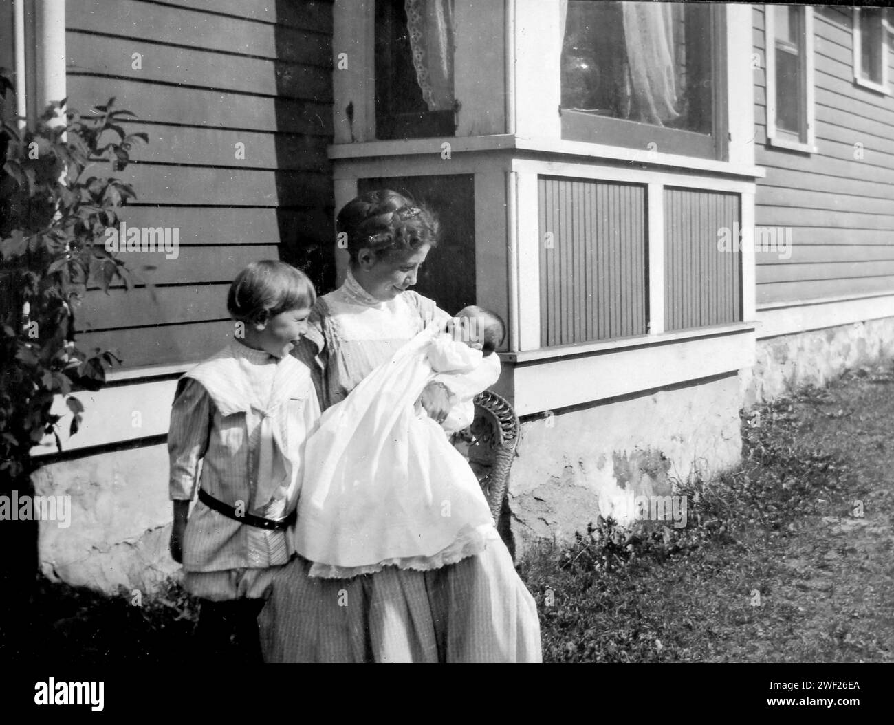 Una giovane madre siede con il suo nuovo nato in un abito da battesimo mentre suo figlio guarda, CA. 1900. Foto Stock