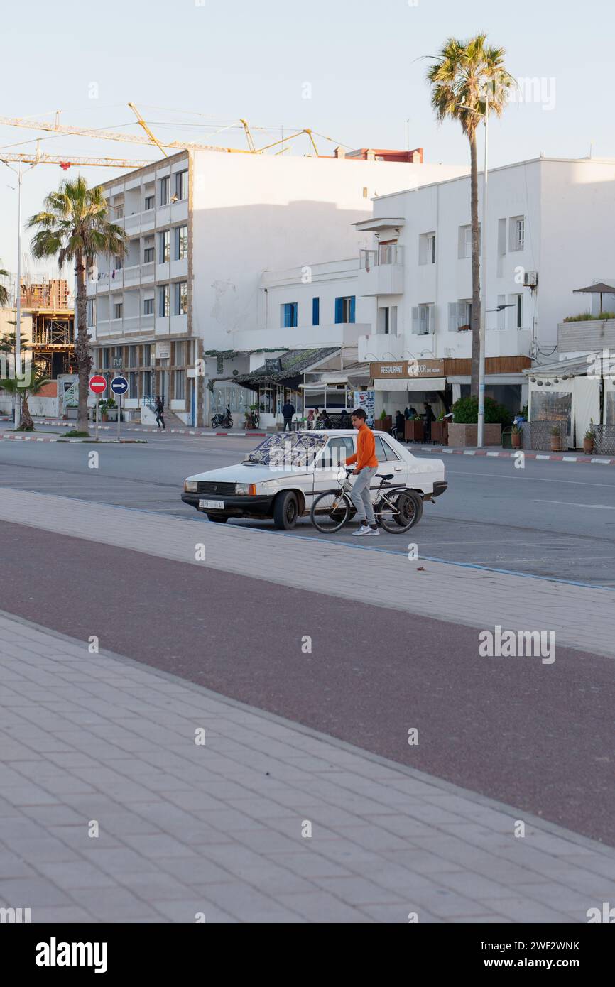 Auto bianca parcheggiata con copertura sul parabrezza con edifici e palme dietro a Essaouira, Marocco, 28 gennaio 2024 Foto Stock