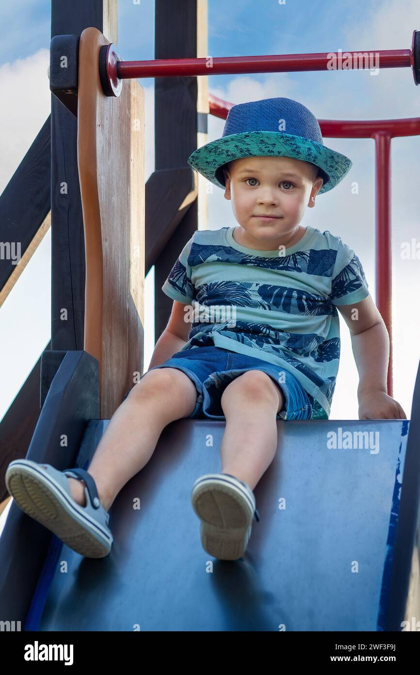 Un bambino felice che si diverte e si arrampica sul parco giochi all'aperto. Simpatico bambino che sorride e si fa strada sullo scivolo. Vacanze estive e primaverili per bambini attivi. Ragazzo Foto Stock
