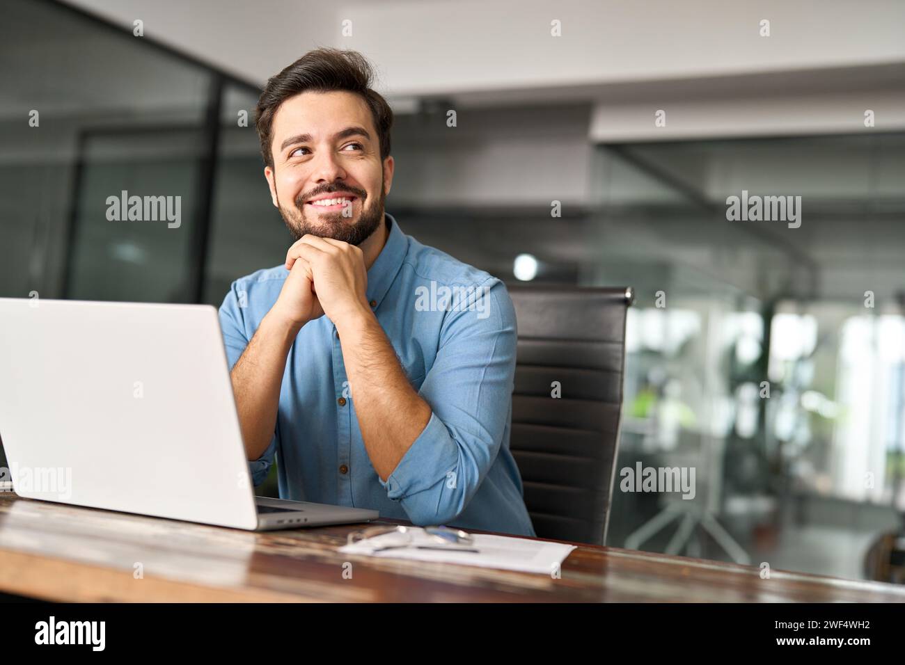 Felice giovane uomo d'affari che lavora su un computer portatile e guarda lontano dalla scrivania dell'ufficio. Foto Stock