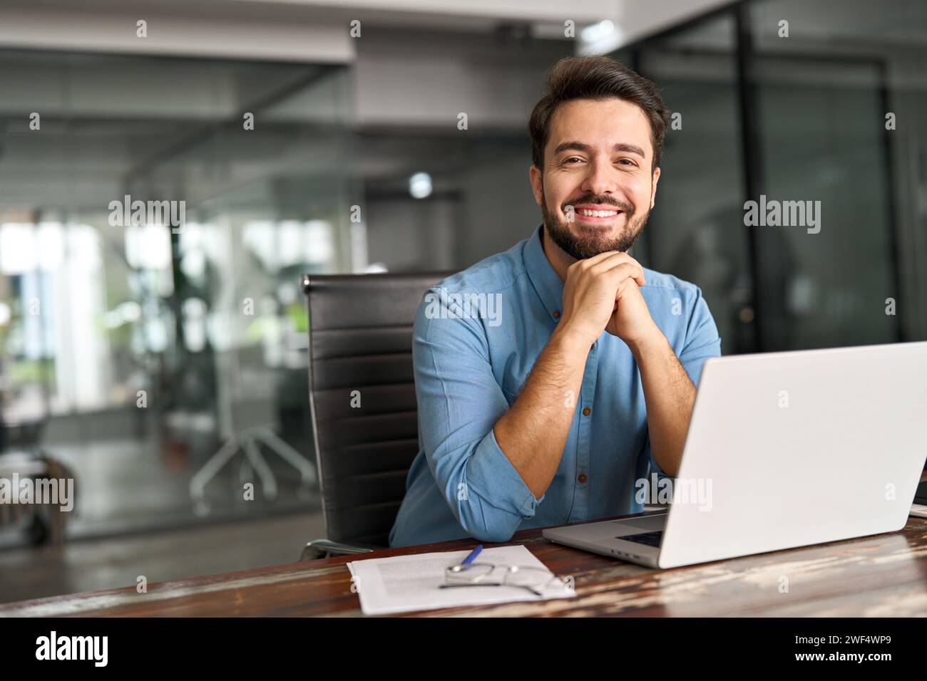 Un giovane uomo d'affari felice che lavora su un computer portatile che guarda la fotocamera alla scrivania dell'ufficio. Foto Stock