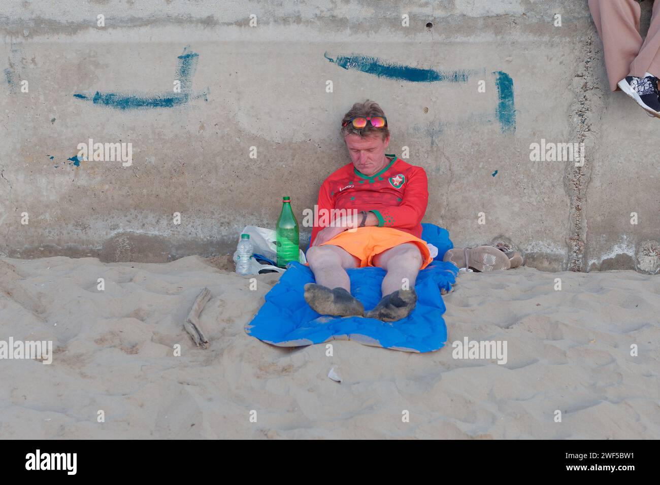 Il turista inglese in maglietta rossa e pantaloncini arancioni e occhiali da sole si addormenta su una spiaggia sabbiosa di Essaouira, Marocco, 28 gennaio 2024 Foto Stock