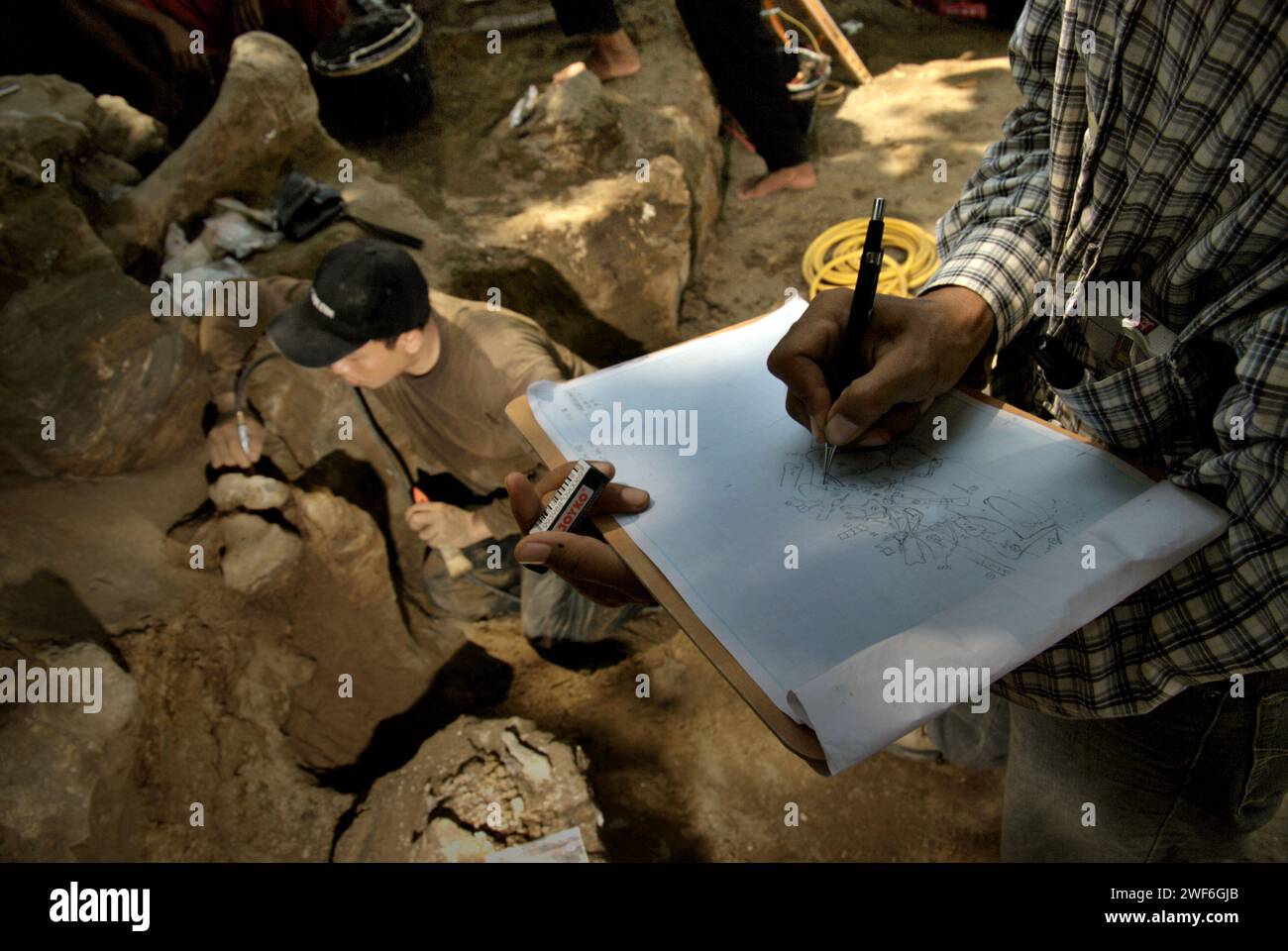 Il paleontologo Iwan Kurniawan disegna uno schizzo su carta che mostra le posizioni delle ossa fossilizzate di Elephas hysudrindicus, una specie di elefante estinta vissuta durante l'epoca del Pleistocene, che in seguito è conosciuta come "elefante Blora" mentre il suo team sta lavorando in background nel sito di scavo di Sunggun, Mendalem, Kradenan, Blora, Central Java, Indonesia. Il team di ricerca sui vertebrati (Agenzia geologica, Ministero dell'energia e delle risorse minerarie indonesiano) guidato dallo stesso Kurniawan con Fachroel Aziz ha scoperto le ossa della specie quasi interamente (circa il 90% completo). Foto Stock